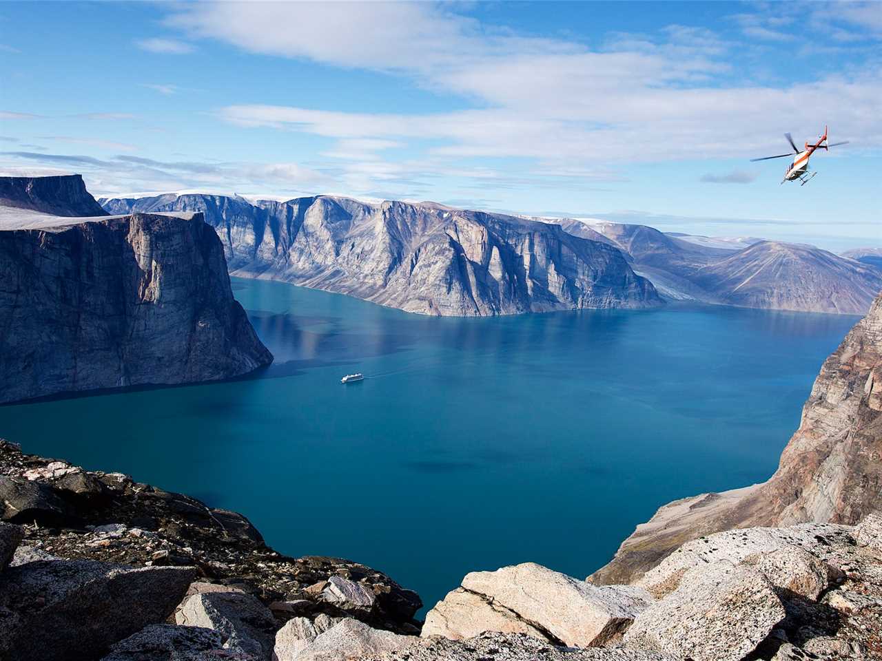 A far shot of The World sailing on water in a mountainous landscape as a helicopter flies overhead.