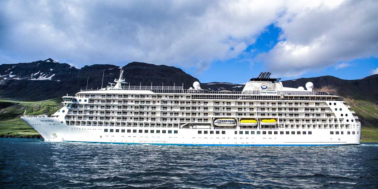 The World sailing past a green, tree-lined coastline.