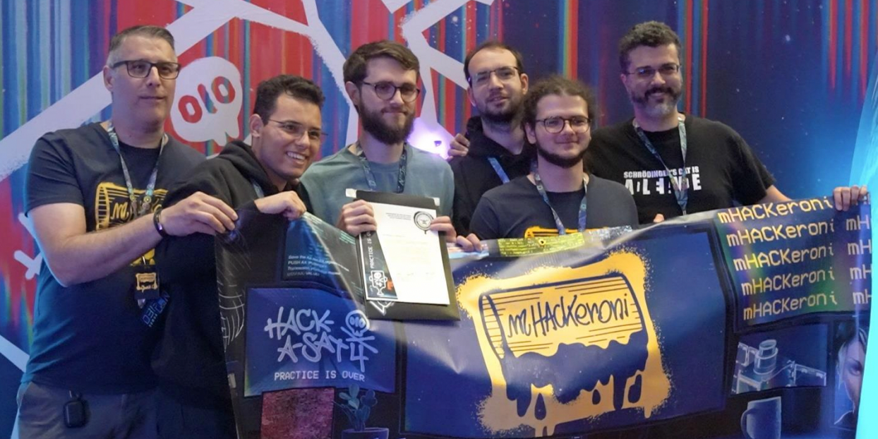 A picture of the Hack-A-Sat winning team, mHACKeroni, shows six people smiling as they hold up flags and a prize.