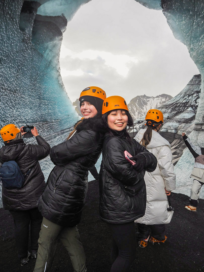 two students in safety helmets pose back to back during a hike in Inceland