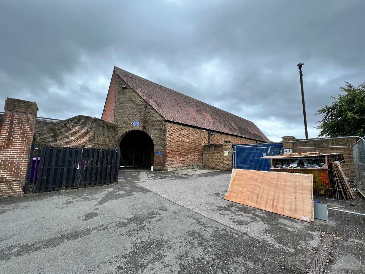 A large windowless brick building with a pointy roof, a converted barn, with a skip and in the front.