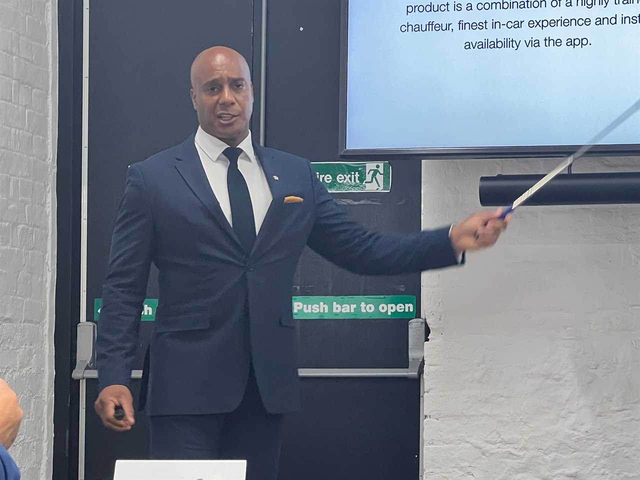 Ricky Miller of Wheely wears a navy suit and points with a stick at a slideshow on a TV