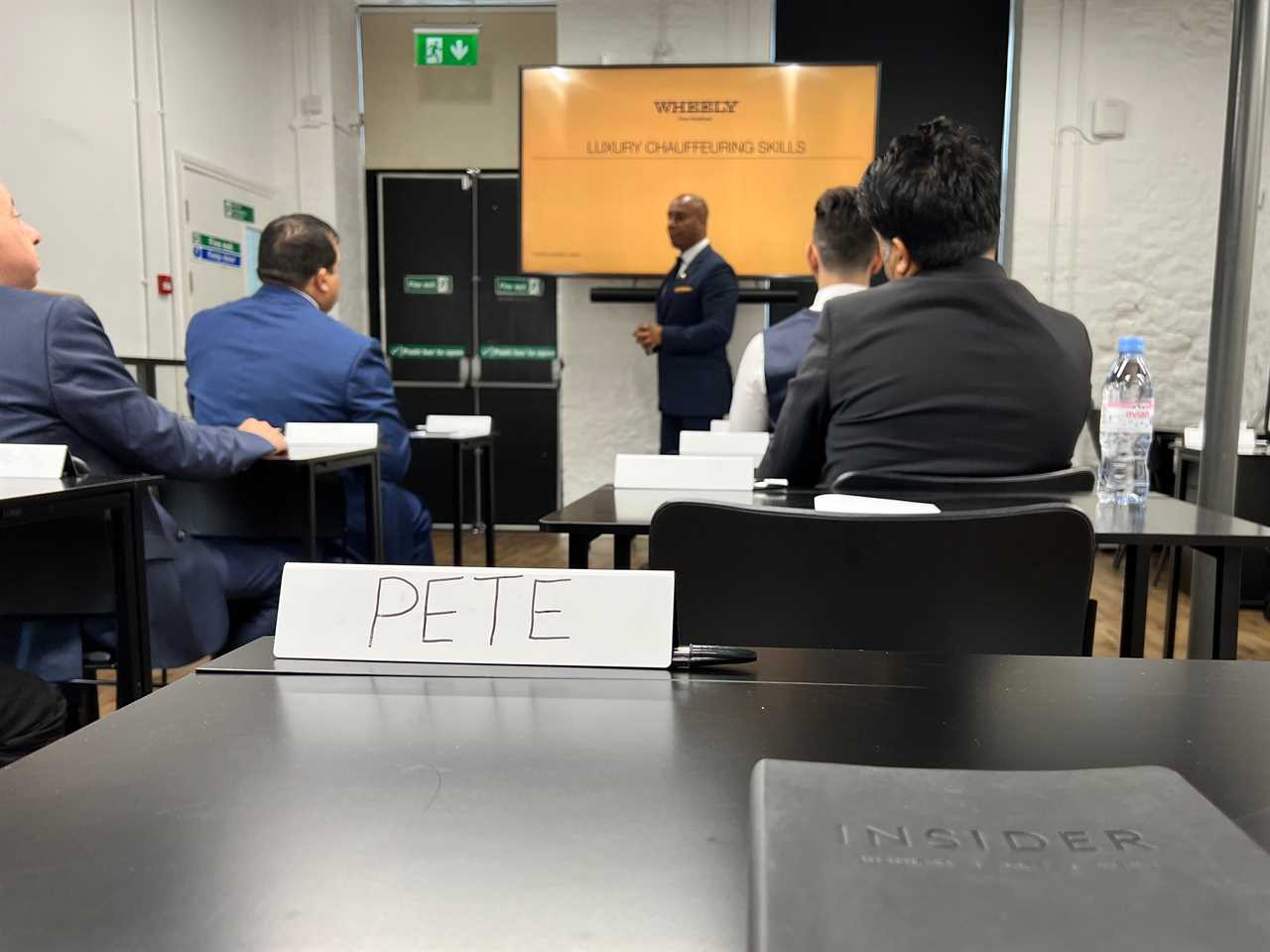 A classroom, four other men in suits are sat down with a teacher at the front. A name tag on the desk says "Pete" with an Insider notebook