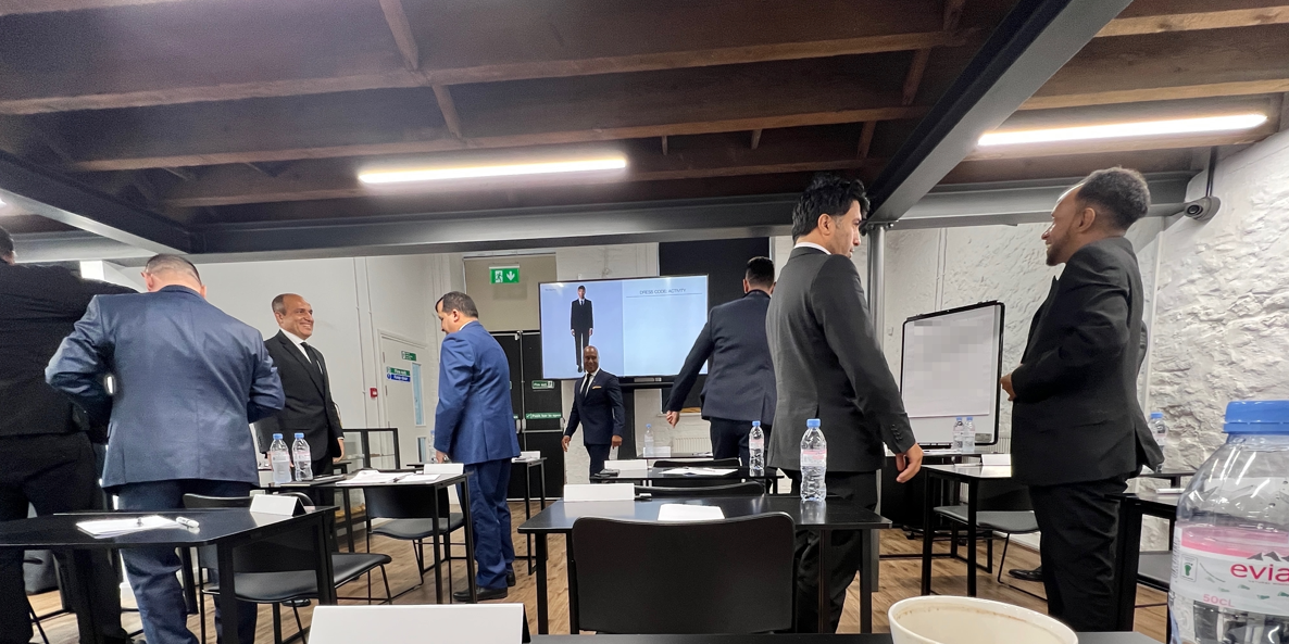 Three pairs of besuited men stand up in a classroom, looking at each other's outfits