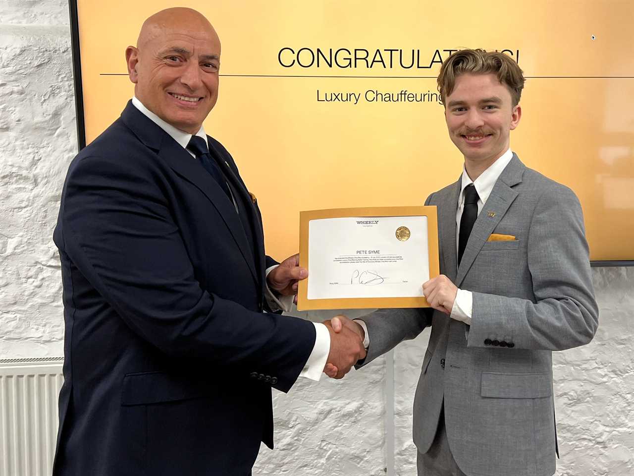 A man in a navy suit and a man in a grey suit shake hands as a certificate is presented and a screen in the background says congratulations