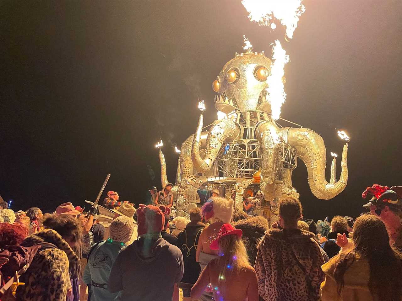 A crowd of dancers at Burning Man festival, surrounding a metal sculpture of an octopus shooting flames mounted on a car