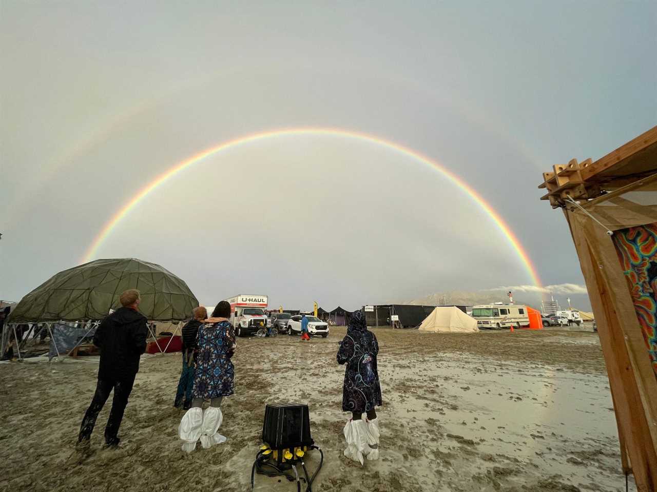 Three people at Burning Man festival in Nevada stand in mud and look at a double rainbow in the sky.