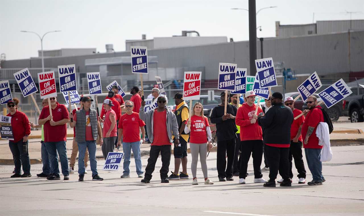 People on strike in red t-shirts carrying blue UAW signs are gathered outside of a Ford vehicle assembly plant. 