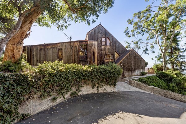 A long, paved drive leads up to the elevated, wood-clad home, surrounded by tall trees and lush vegetation. The lower level houses a spacious five-car garage.