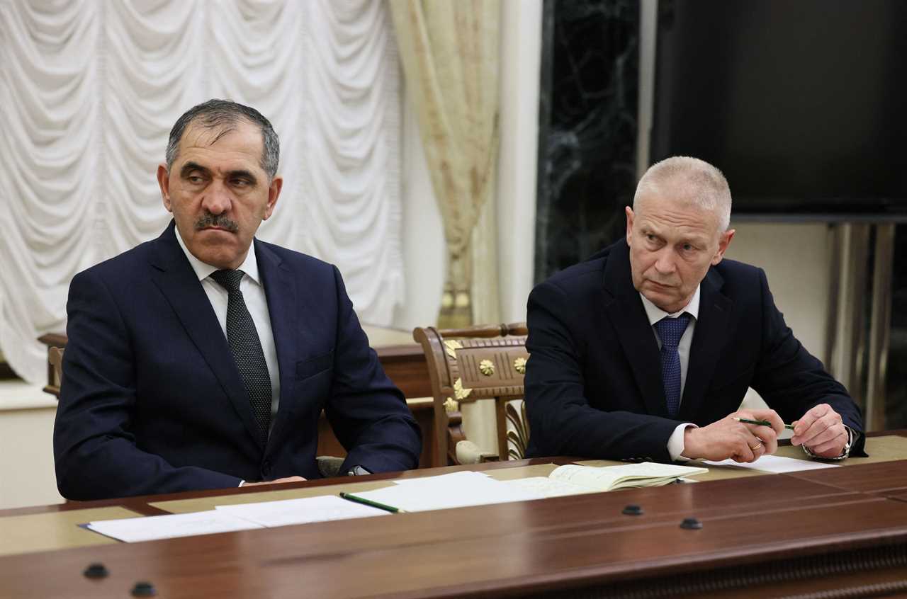 two old white men in suits seated at wooden table looking grumpy