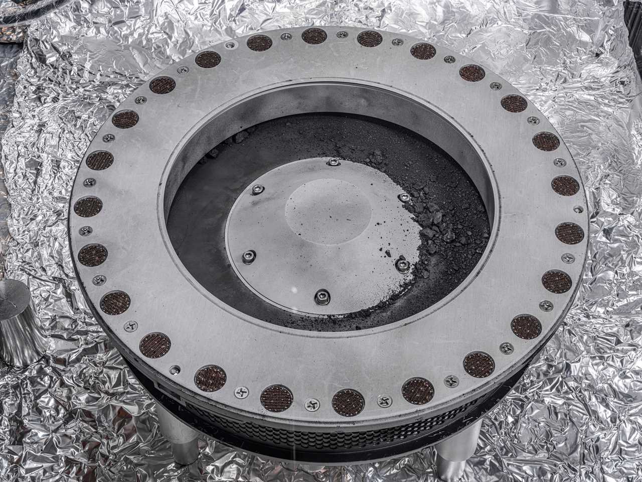 A circular piece of the container holding the Bennu asteroid sample with black rocks from the asteroid on it