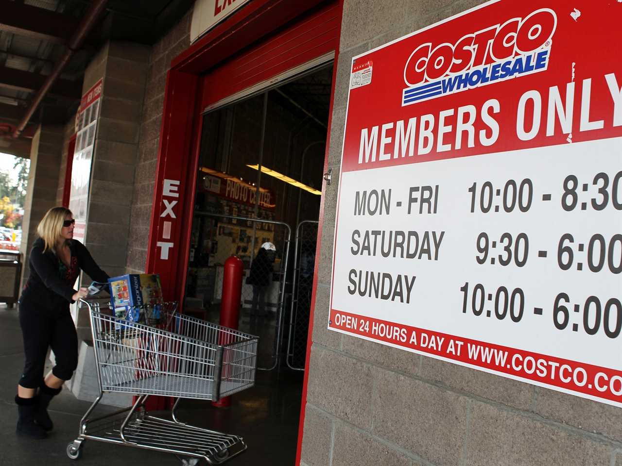 A shopper pushes a cart into a Costco store next to sign displaying hours