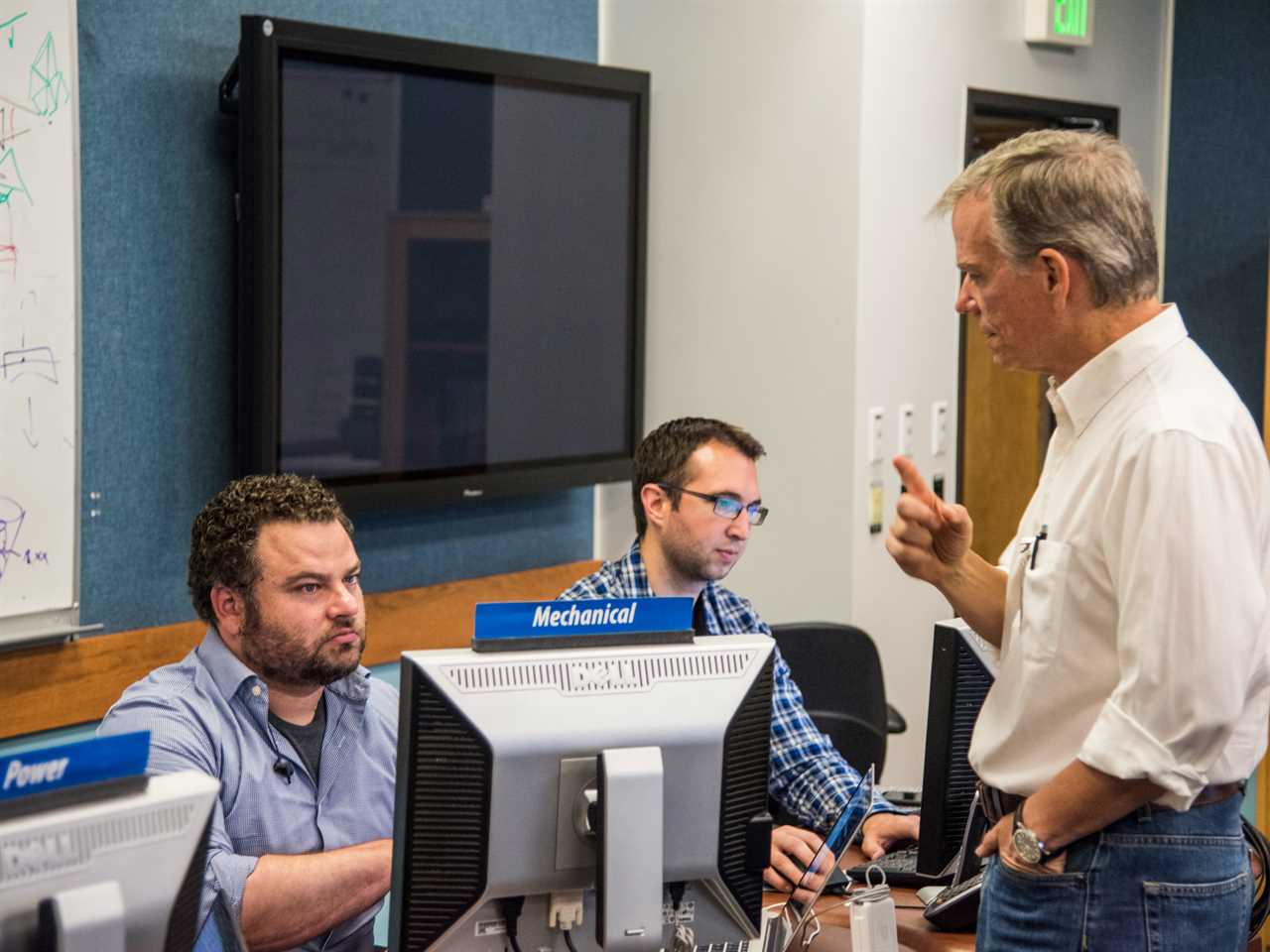 a man stands talking to two colleagues over their monitors