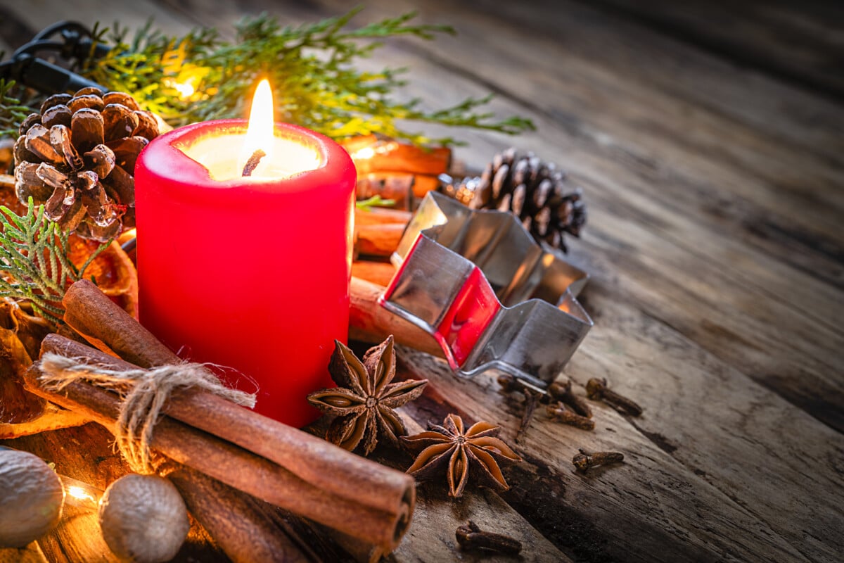 Christmas backgrounds: red burning Christmas candle shot on rustic wooden table. Cinnamon sticks, star anise, cloves and nutmeg are around the candle. The composition is at the left of an horizontal frame leaving useful copy space for text and/or logo at the right. Predominant colors are red and brown.