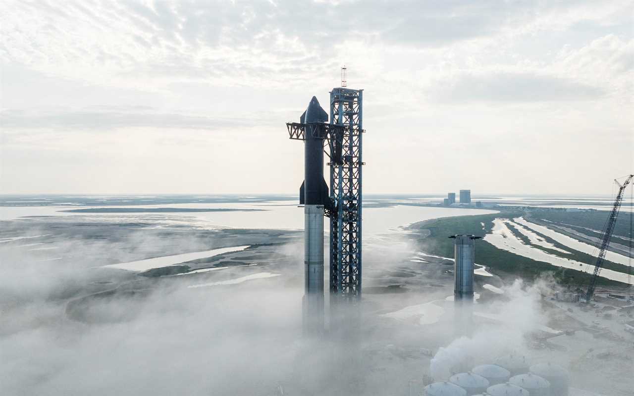 starship black rocket on silver booster secured to a launch tower above foggy flat wetlands