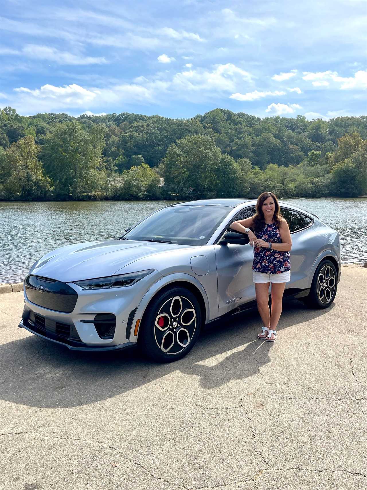 A woman stands in front of her silver SUV in front of a lake with green trees.