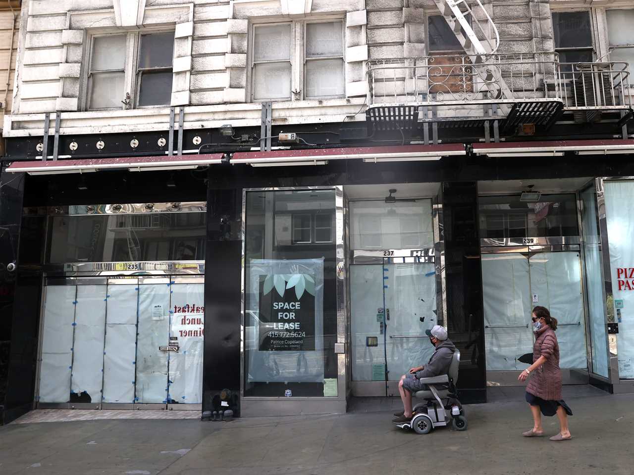 Multiple vacant storefronts on a city street with papered-over windows and a couple people walking past