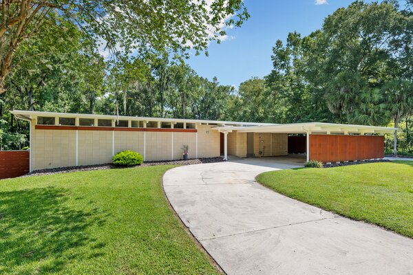 The front of the home features a large covered carport, accessed via the paved, U-shaped driveway. Soaring trees surround the property, enhancing the overall sense of privacy.