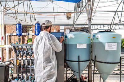 Christian Bake, wearing a long white coat, standing in front of an automated irrigation system inside a greenhouse.