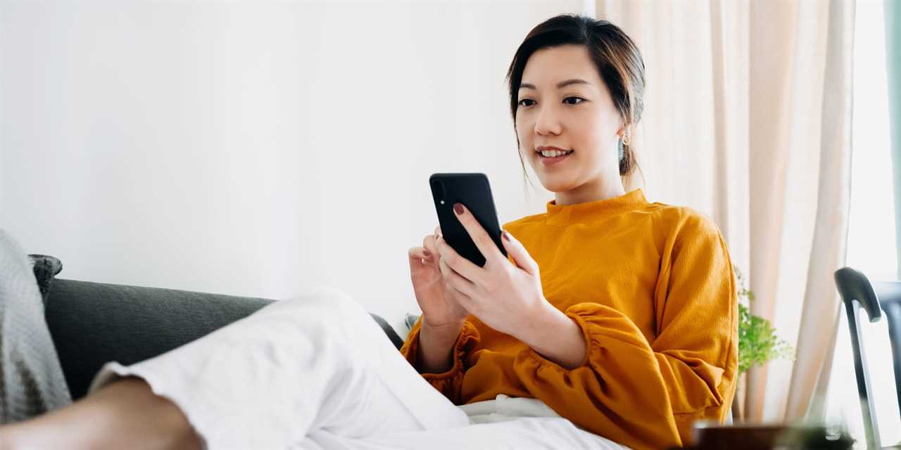 woman in yellow sweater and white pants laying on a coach and looking at phone.