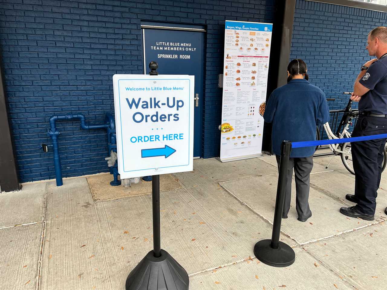 A sign says "Walk-up orders" and points customers toward a line where a Little Blue Menu employee stands with a tablet taking a customer's order as he looks at a poster with the restaurant's menu. In the background, signage on a door reads: "Little Blue Menu team members only."