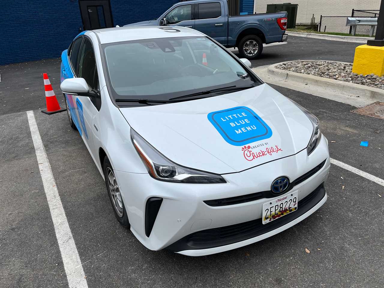 A Little Blue Menu Toyota Prius delivery car sits in a parking spot outside the restaurant.