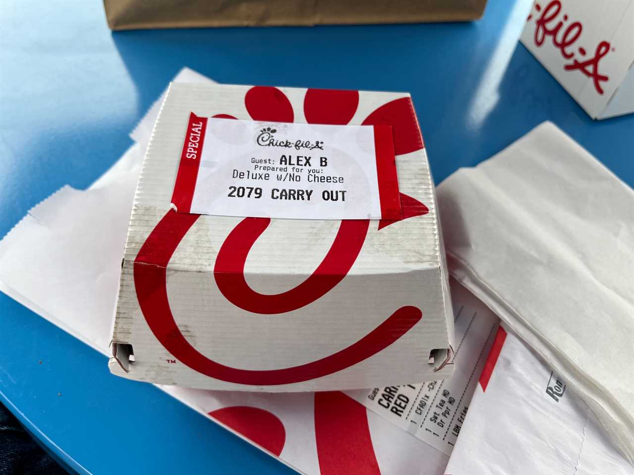 A Chick-fil-A chicken sandwich sits in a red-and-white cardboard box with the restaurant's logo and a label with the reporter's name and "Carry out" at Little Blue Menu