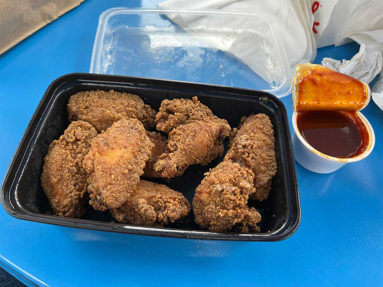 Chicken wings sit in a plastic container at Little Blue Menu with a cup of sauce next to them