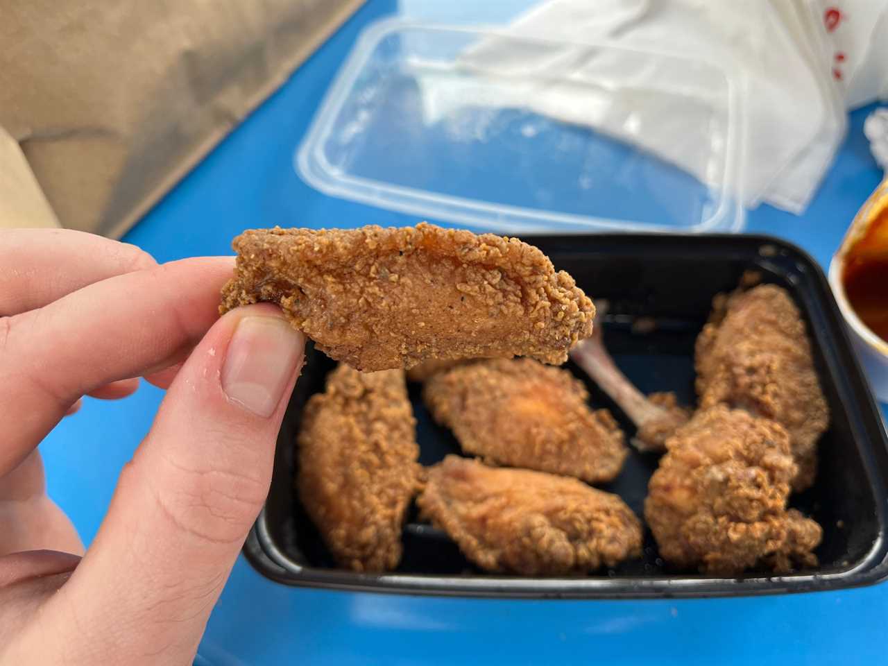 The reporter holds up a chicken wing above a plastic container of chicken wings sitting on a blue table at Little Blue Menu