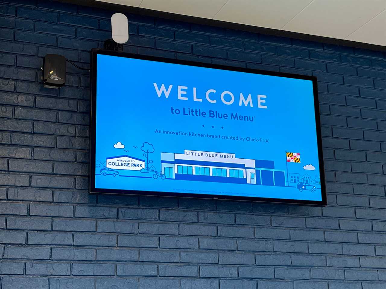 A screen near the front of Little Blue Menu reads "Welcome to Little Blue Menu: An innovation kitchen brand created by Chick-fil-A" and includes a cartoon illustration of the restaurant and the flag of the state of Maryland.