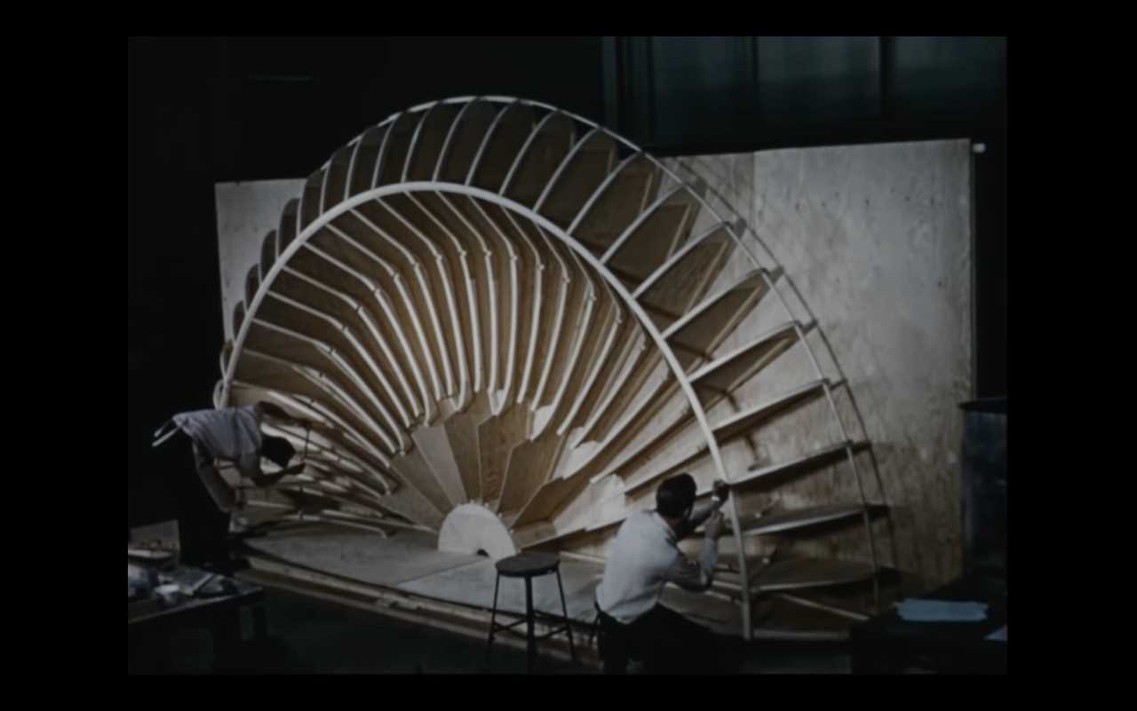 Two men stand on opposite sides of a wooden half-circle that would become the inner skeleton of the to-scale model of the Avrocar.