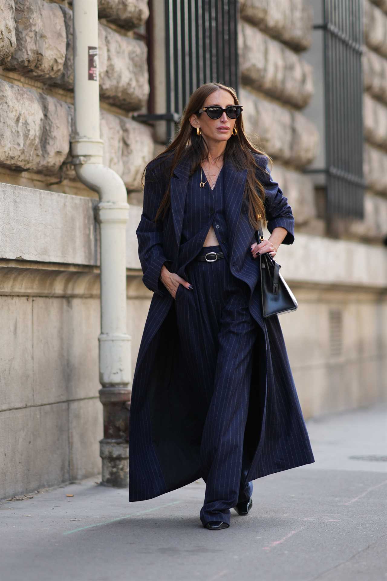 A women walking down the street in Paris wearing a pinstripe shirt, pants and trench coat with black sunglasses and chunky gold earrings