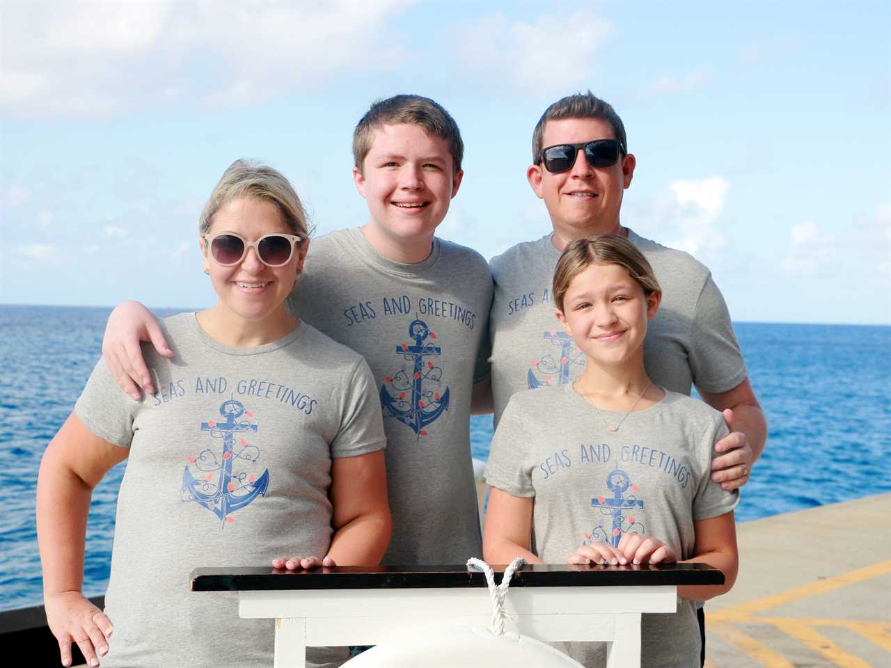 Terri Peters and her family wearing grey t-shirts that say "Seas And Greetings" with a blue anchor wrapped in red Christmas lights. Terri wears pink sunglasses and has dyed blonde hair. Her son has dark brown hair and has his right arm around her shoulders. His other arm wraps around his father, to his right. His father has black sunglasses on and dark brown hair gelled up. He has his arm around his son and his left around his daughter's shoulder. She stands in front of him and has blonde hair tied back.