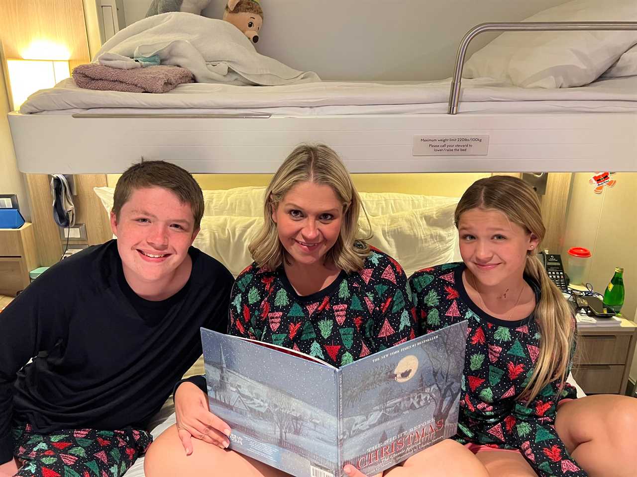 Terri Peters reading "Twas the Night Before Christmas" with her son on her right side and her daughter on her left. They smile at the camera, while wearing matching Christmas pajamas. The pjs are black with a pattern of pink, green, and red evergreen trees and white dots like snow. The son wears a black long-sleeved shirt but the pants match the other two's pjs.