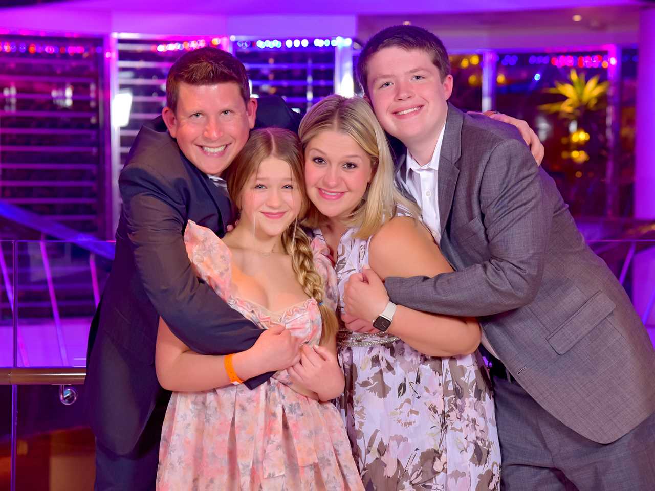 Photo of Terri and her family posing and hugging. Terri's husband stands on the left with his arm around his daughter in front of him and his son to his right. He wears a navy suit. Terri's daughter is in front of him and wears a flouncy floral dress with puffy sleeves and her hair in a braid, hanging over her left shoulder. She has both arms around her dad's arm which wraps her waist. Terri stands to her right and wears a brown and white patterned sleeveless dress. Her son stands to her right and wraps his arm around her waist, with his other around his dad's.  Terri holds her son's arm. Her son wears a grey suit.