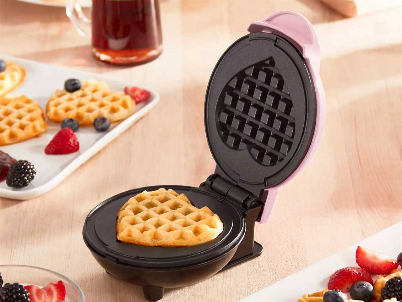 Dash mini waffle maker open to show a heart-shaped waffle inside set on a counter with plates filled with waffles and fruit.