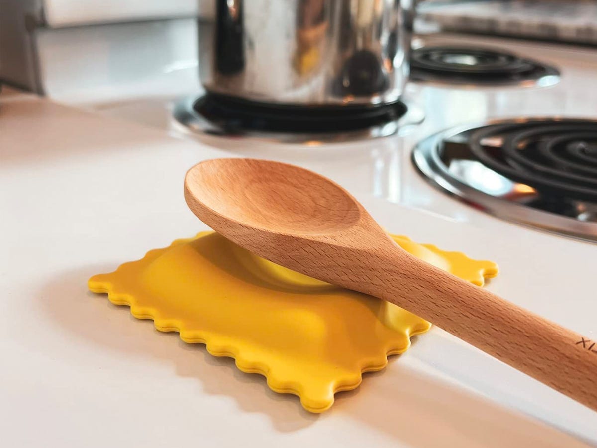 wooden spoon resting on the Genuine Fred Sauced Up ravioli-shaped spoon rest set on a kitchen counter by a stovetop.