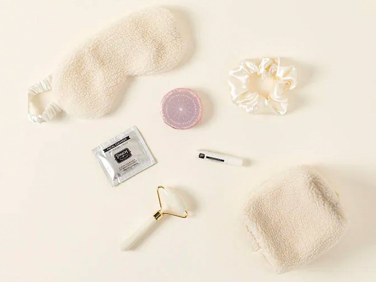 Contents of the Uncommon Goods Wind Down and Relax Kit, including a silk scrunchie, cooling facial towelette, facial roller, lavender shower steamer, bookmark, eye mask, and a zippered pouch