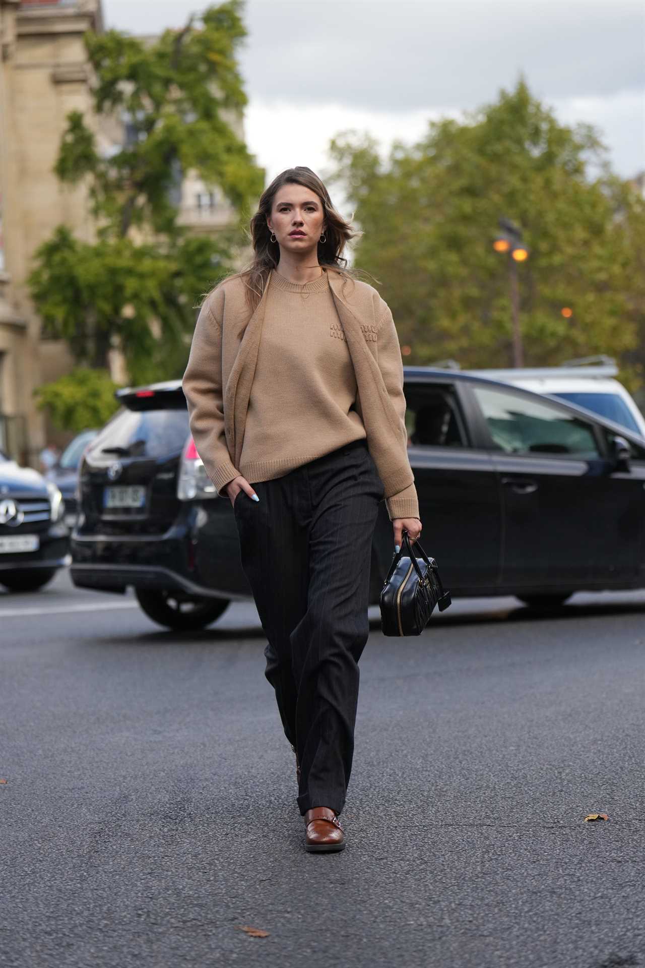 A woman walking on the street in paris wearing a beige sweater and jacket, black pants and brown shoes