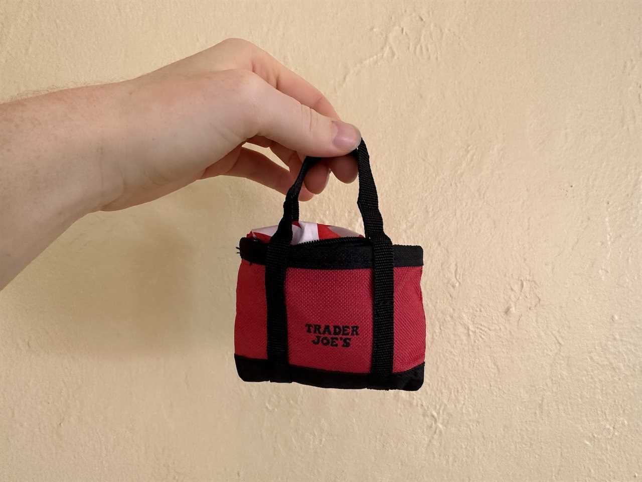 hand holding miniature red and black tote bag that says trader joe's against a beige background