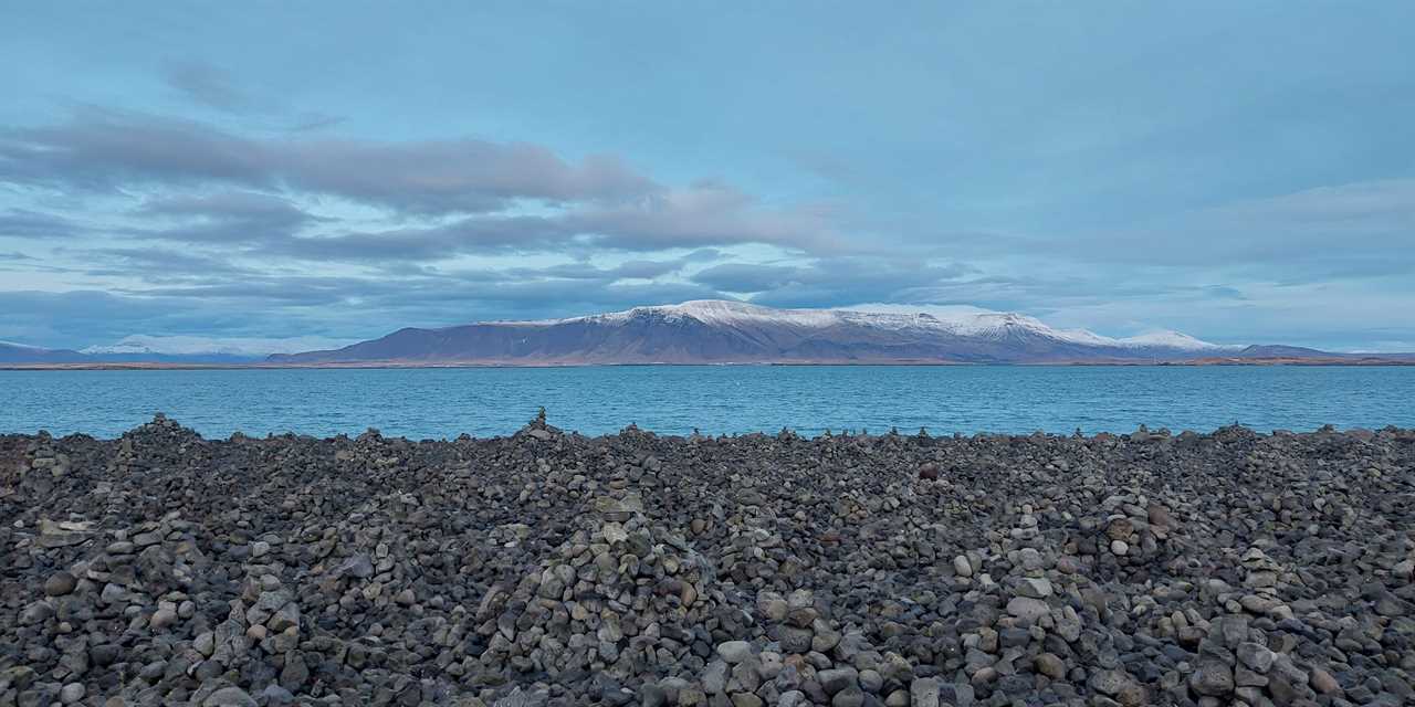 The sea near Reykjavik. Photo shows blue sea, stones on the beach, blue sky and mountains in the distance.