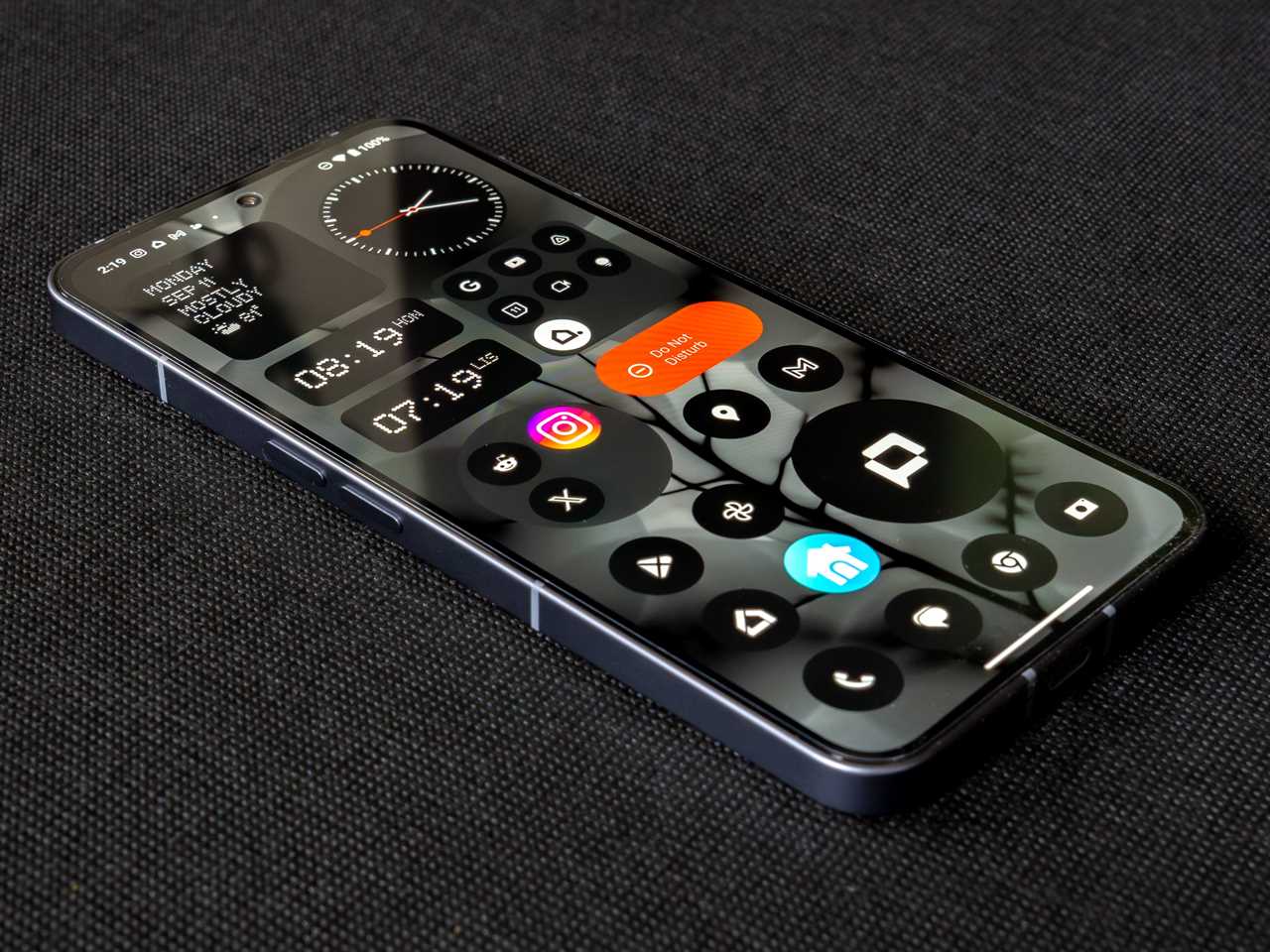 The Nothing Phone laying on a black fabric background at an angle, showing its left edge and display on the home screen.