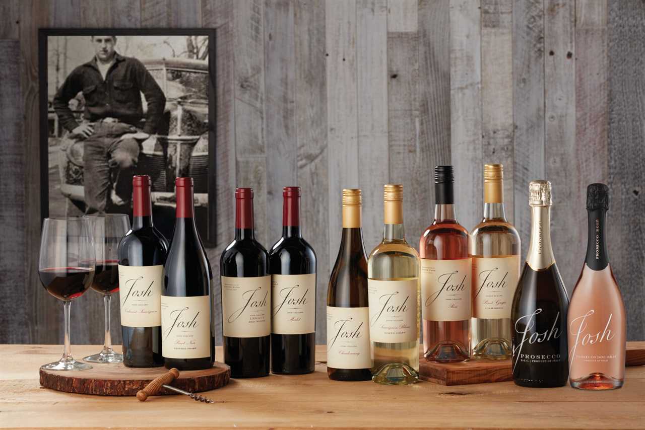 A selection of bottles of wine from Josh Cellars, including red, white, and roses, sits on a wooden-top table with two glasses of red wine and a corkscrew. In the background, a black-and-white photo of Josh, Joseph Carr's father, hangs on a wooden wall.