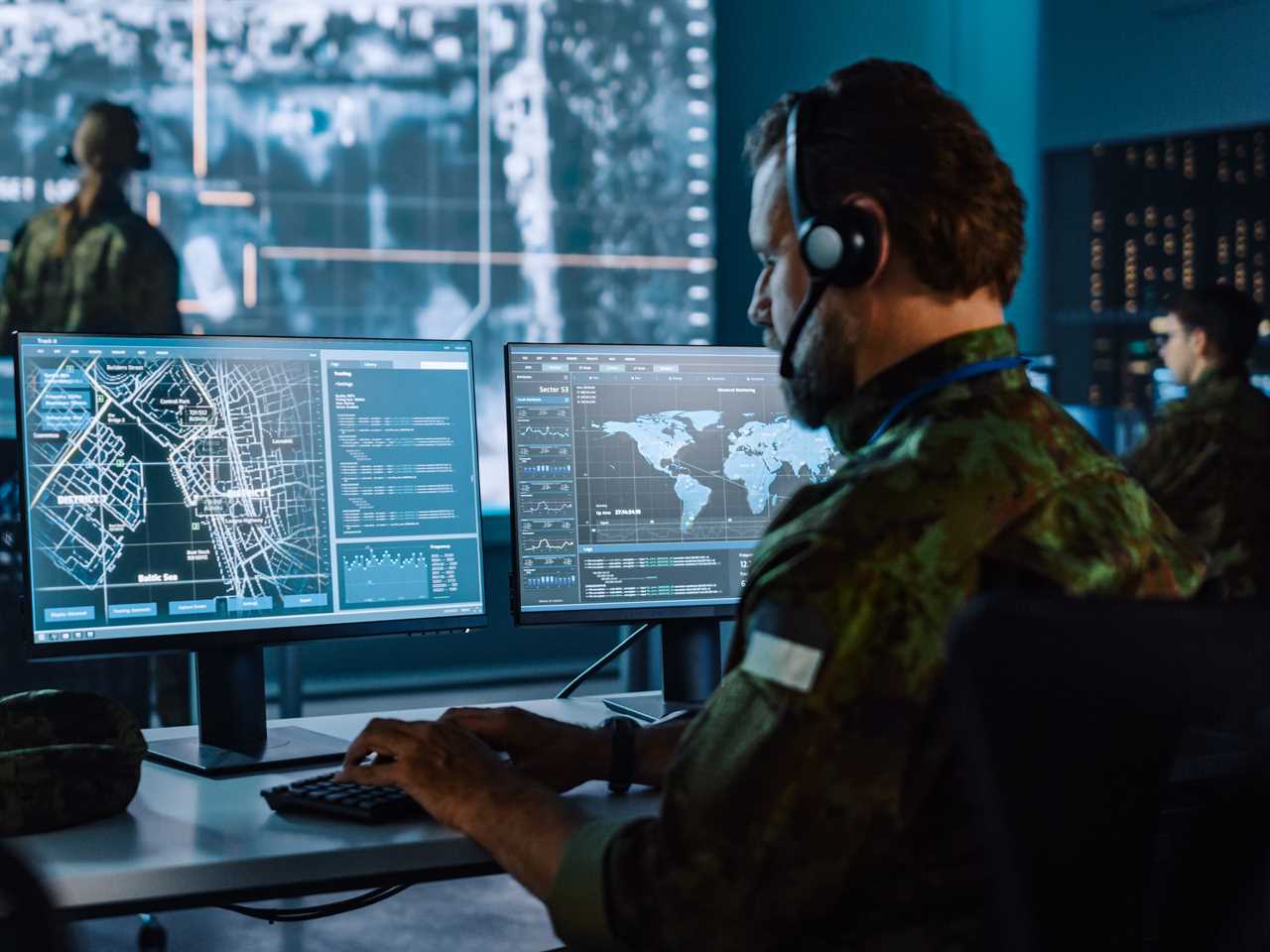 A military officer working on a city tracking operation, sitting at a desk with a headphone microphone and wearing a camouflage shirt.