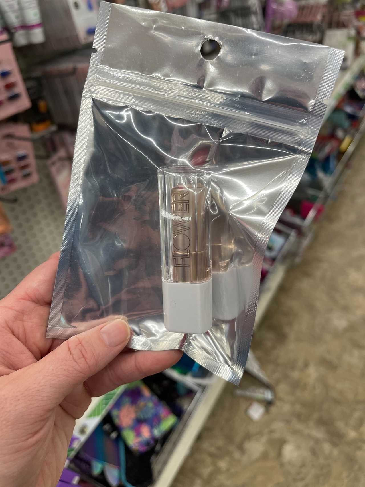 The photographer holds a silver-colored plastic zip lock bag with a tube of lipstick from Drew Barrymore's Flower brand while standing in the aisle of a Dollar Tree store in Ohio.