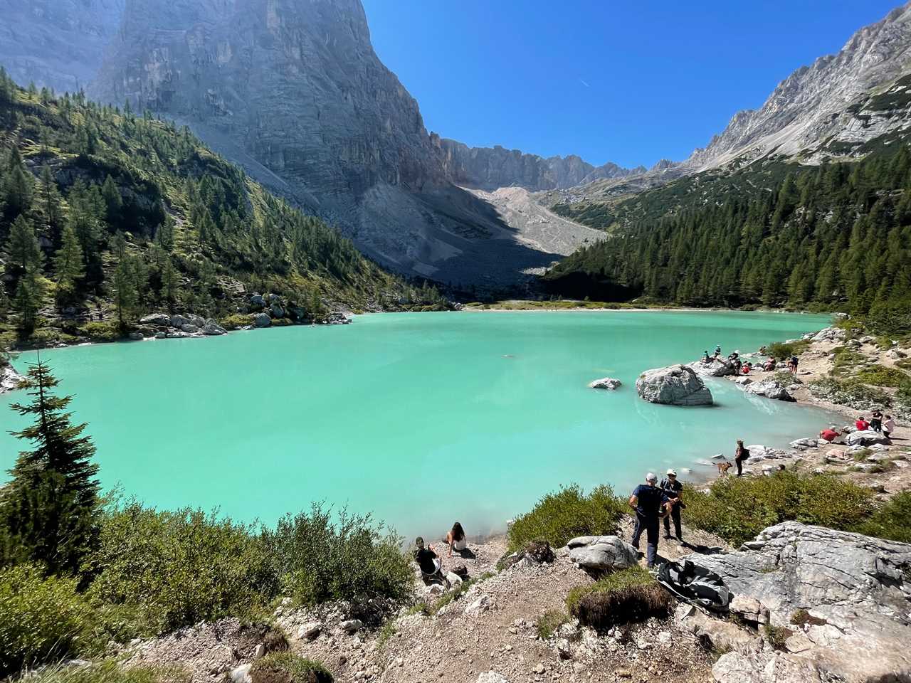 A bright-blue lagoon surrounded by rocks, mountains, and pine trees. People sitting and standing around the rocks surround water