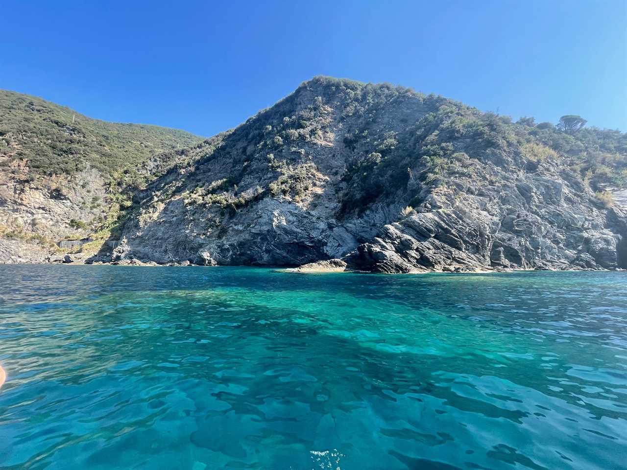 Bright-blue water leading to rocky, tree-filled cliffs with bright-blue skies in Italy