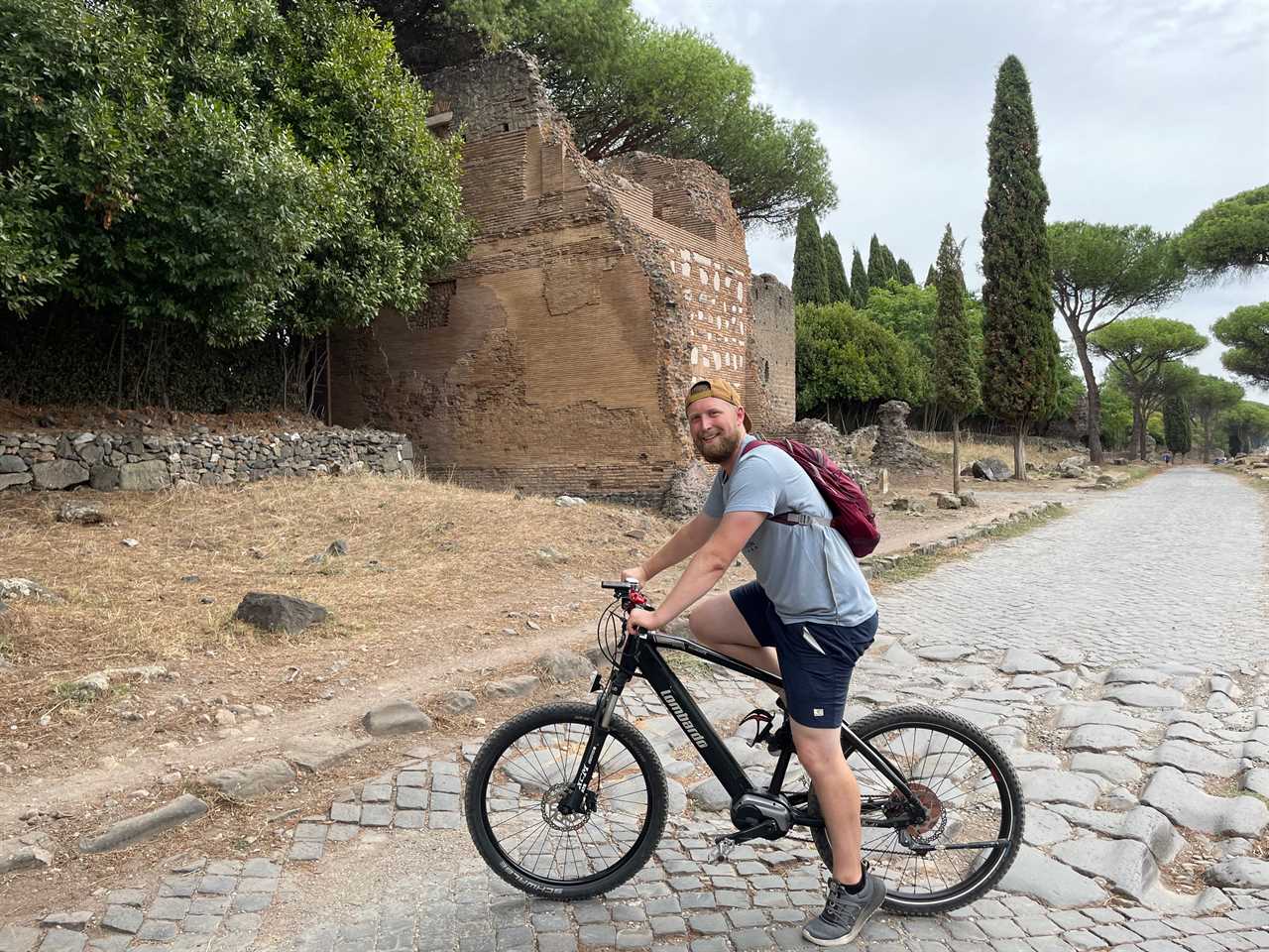 The writer, wearing blue shorts and a T-shirt and red backpack rides a black bike in front of ruins along the Appian Way