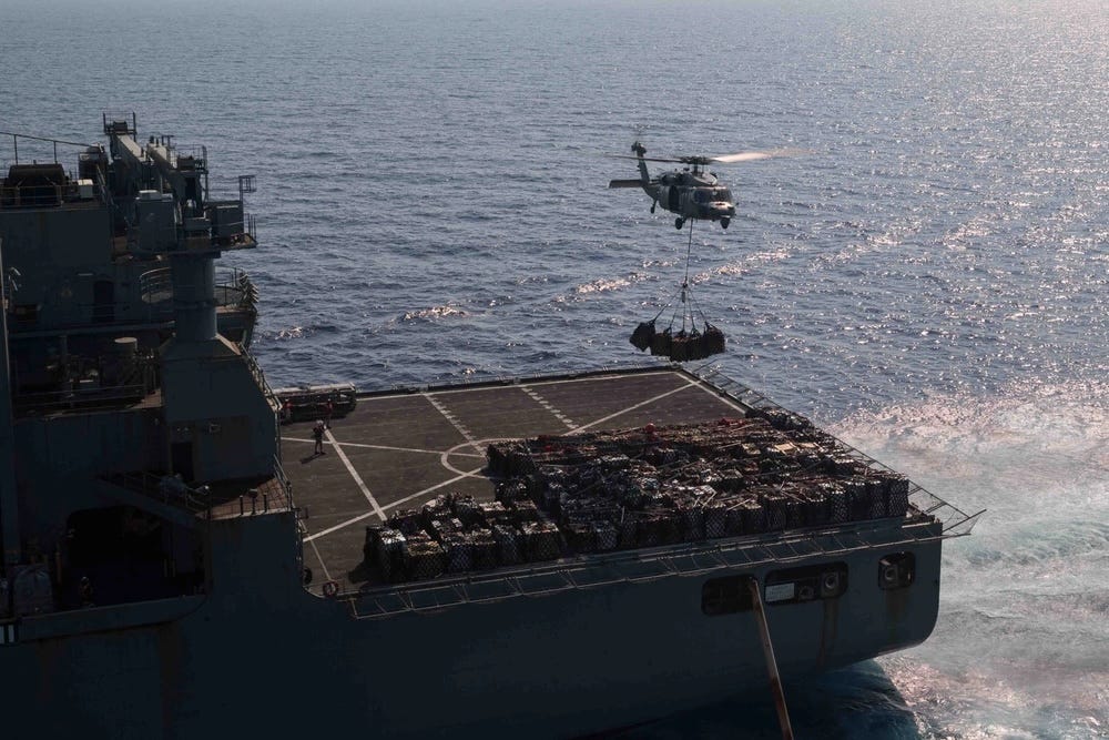 a helicopter carrying cargo hovers above the flight deck of a military ship