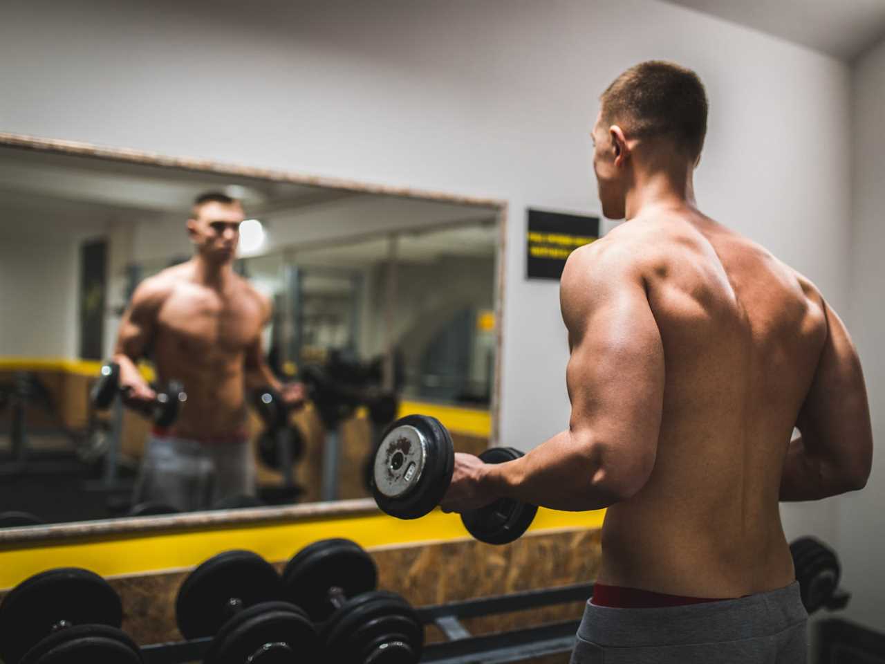 a man whose face isn't visible is lifting weights shirtless in gray sweatpants while looking in a mirror